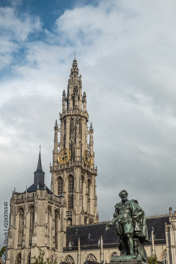 Antwerp, Belgium - September 24, 2018: Peter Paul Rubens bronze statue with towers of Onze-Lieve-Vrouwe Cathedral of Our Lady in back under white cloudy sky.