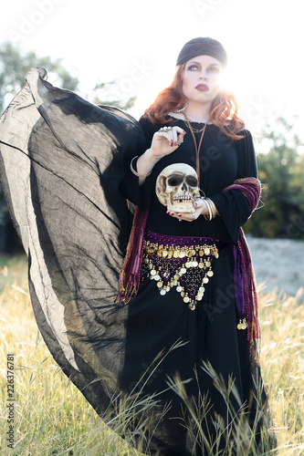 Beautiful young woman with red hair in a witch gypsy or fortune teller costume holding a skull in a meadow field photo