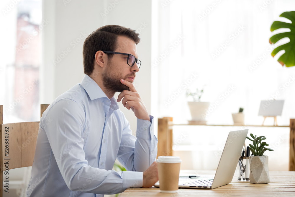 Concentrated thoughtful millennial businessman or business owner with eyeglasses sitting at the desk in office room or home working using computer. Thinking about new project, making decision concept