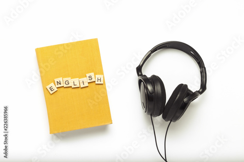 Book with a yellow cover with text English and black headphones on a white background. Concept of audio books, self-education and learning English independently. Flat lay, top view
