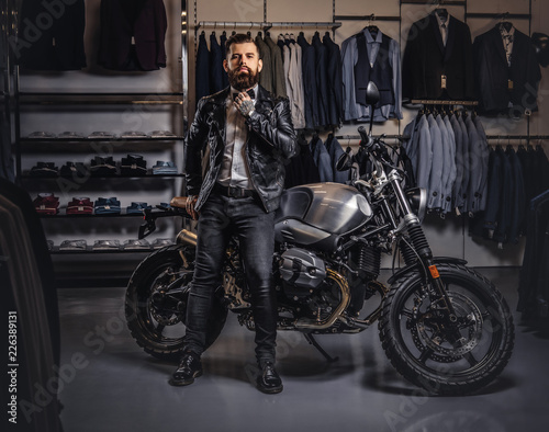 Stylish tattooed bearded man with dressed in black leather jacket and bow tie posing near retro sports motorbike at men's clothing store. © Fxquadro