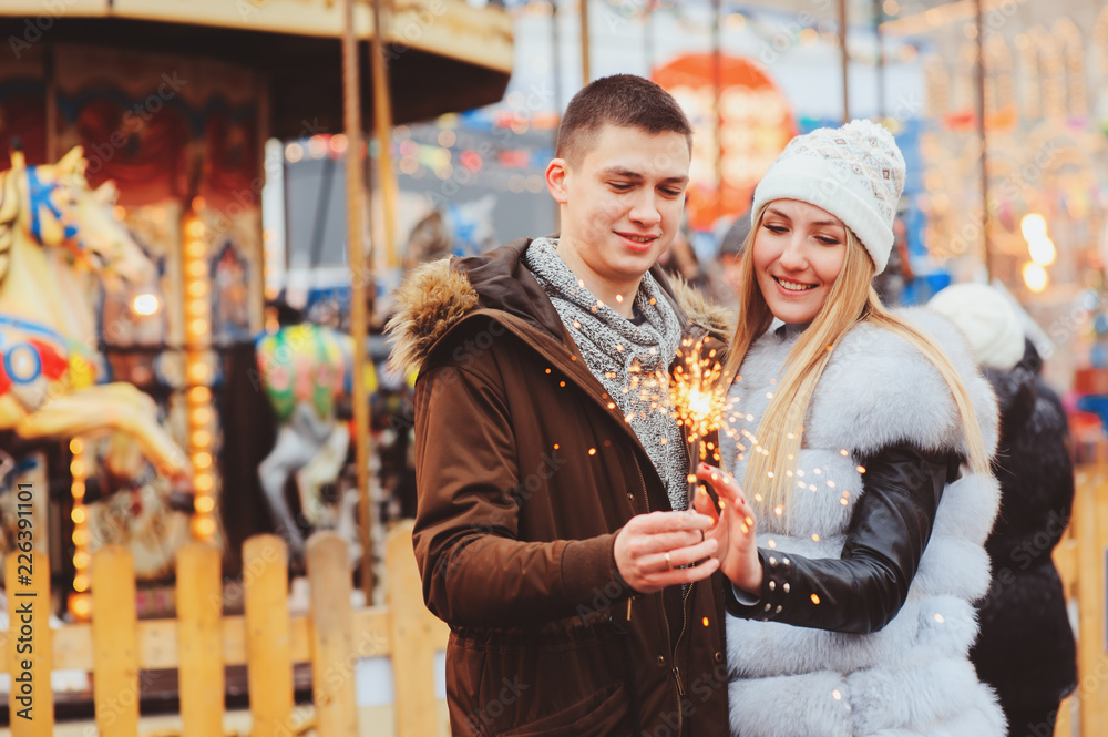happy couple with christmas firelights walking in city holiday amusement park, carousel on background happy couple with christmas firelights walking in city holiday amusement park, carousel on background