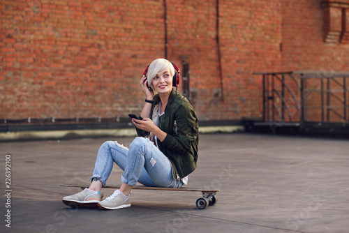 Fototapeta Naklejka Na Ścianę i Meble -  Pretty blond girl listening music in red headphones and using smartphone while sitting on the longboard at the background of brick wall.