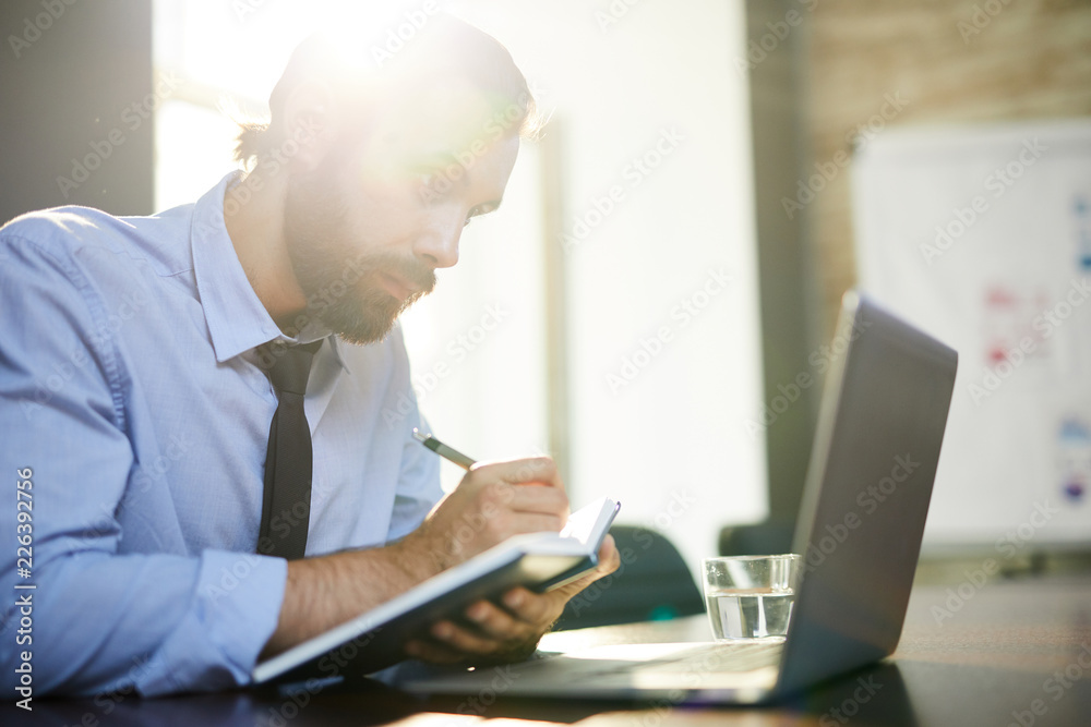 Young man with notebook and pen making notes while looking at laptop display Young man with notebook and pen making notes while looking at laptop display