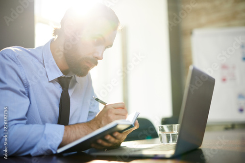Young man with notebook and pen making notes while looking at laptop display