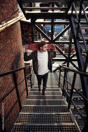Young handsome hipster man with skateboard on the black iron stairs of the industrial building. © gorynvd