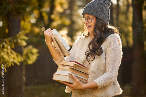 beautiful woman in a cap and glasses is holding a pile of books on the background of autumn forest