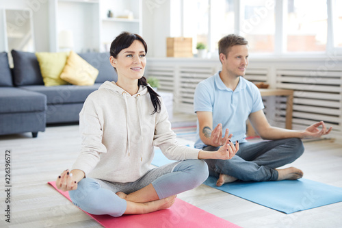 Young active couple sitting in pose of lotus on the floor of living-room during home yoga workout