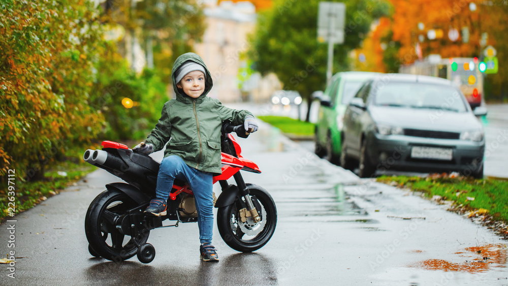 A beautiful little boy posing at his children's red motorcycle. The concept of teaching children to motor vehicles from an early age A beautiful little boy posing at his children's red motorcycle. The concept of teaching children to motor vehicles from an early age