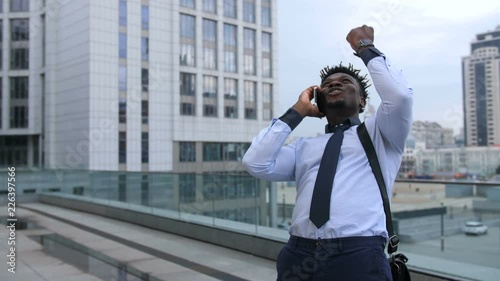 Ecstatic african american executive holding smart phone and celebrating his success after good news with fist clenched. Excited black businessman rejoicing his achievement with winnig gesture outdoors photo