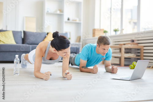 Contemporary young active couple in sportswear doing planks on the floor in front of laptop while watching online workout