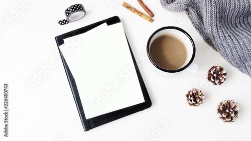 Christmas desktop. Blank greeting card mockup on black notepad, cup of coffee, washi tape, knitted plaid, cinnamon and pine cones on white table background. Winter office, Flat lay, top view. Loopable