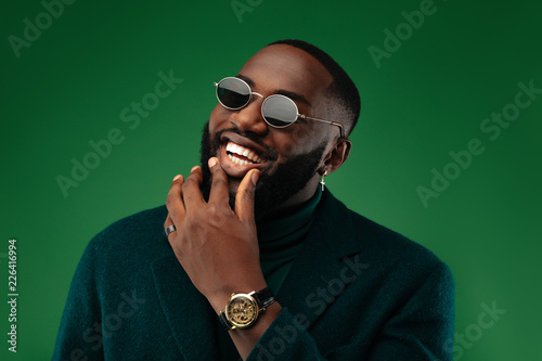 Man portrait. Style. Handsome Afro American guy in green jacket and sun glasses is looking at camera and smiling, on a green background