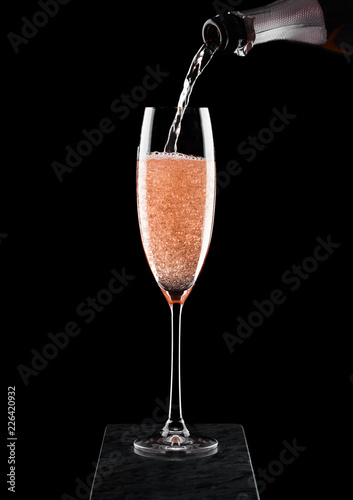 Pouring pink rose champagne from bottle to glass on black marble board on black background. Space for text