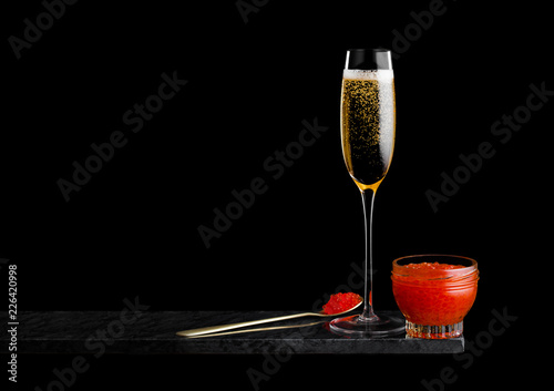 Elegant glass of yellow champagne with red caviar on golden spoon and glass container of caviar on marble board on black background. Space for text