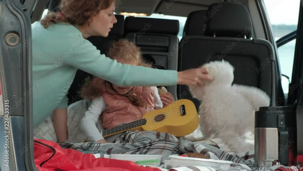 Beautiful little girl sitting in an open trunk of a car on the river bank of the sea playing on a yellow ukulele Beautiful little girl sitting in an open trunk of a car on the river bank of the sea playing on a yellow ukulele