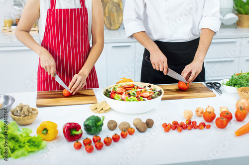 Beautiful Asian woman in red apron learning how to cook and mix vegetable salad in big bowl, teaching by Asian chef in white modern kitchen