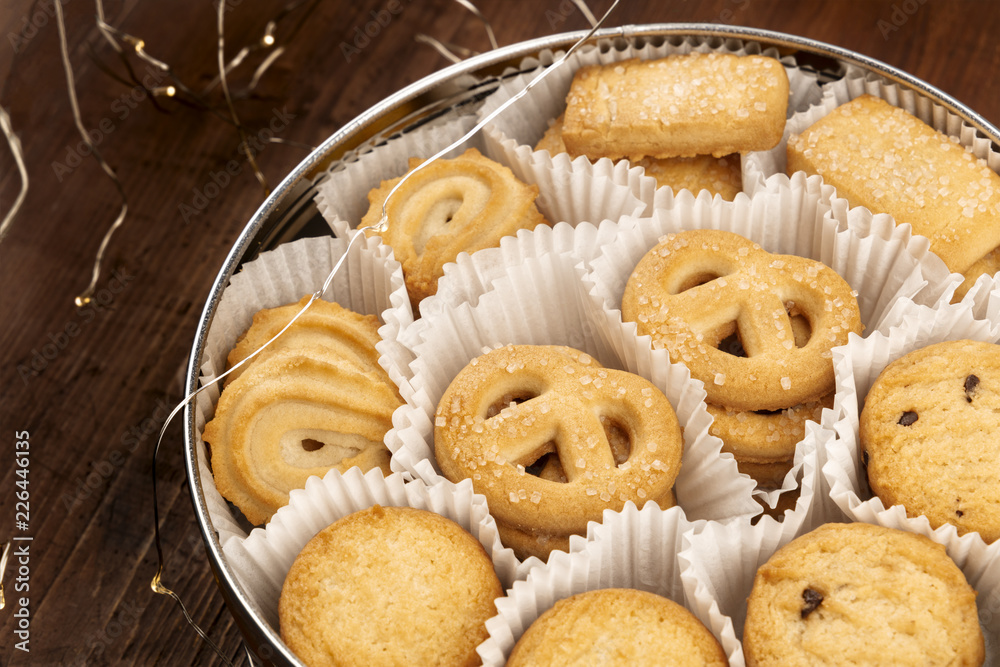 A photo of Danish butter cookies in the traditional tin box, on a dark rustic background with Christmas lights and copy space