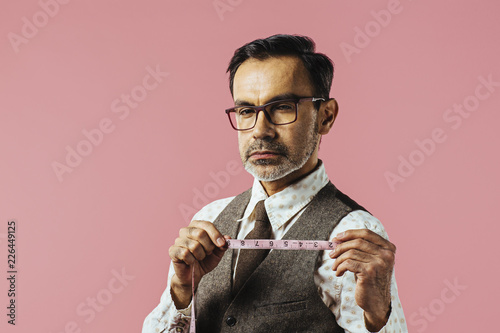 Close up portrait of a tailor holding fabric measure, isolated on pink studio background