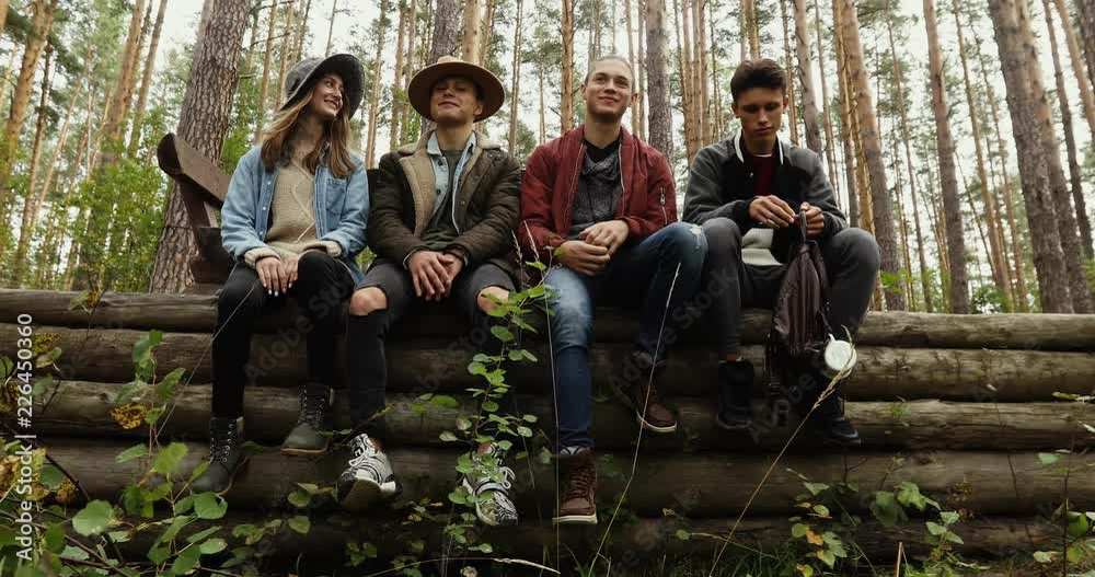 Four young people in jeans and jackets sitting on pile of logs in forest on background of trees. One guy is sad he is offended Four young people in jeans and jackets sitting on pile of logs in forest on background of trees. One guy is sad he is offended