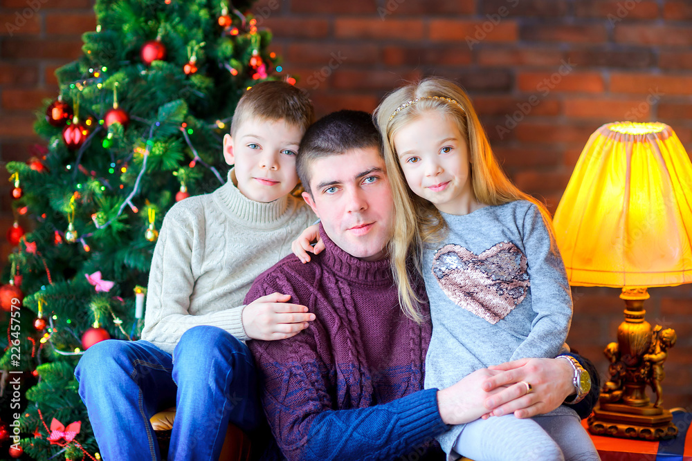 dad with children sitting in a chair near the Christmas tree, gently hugging and smiling