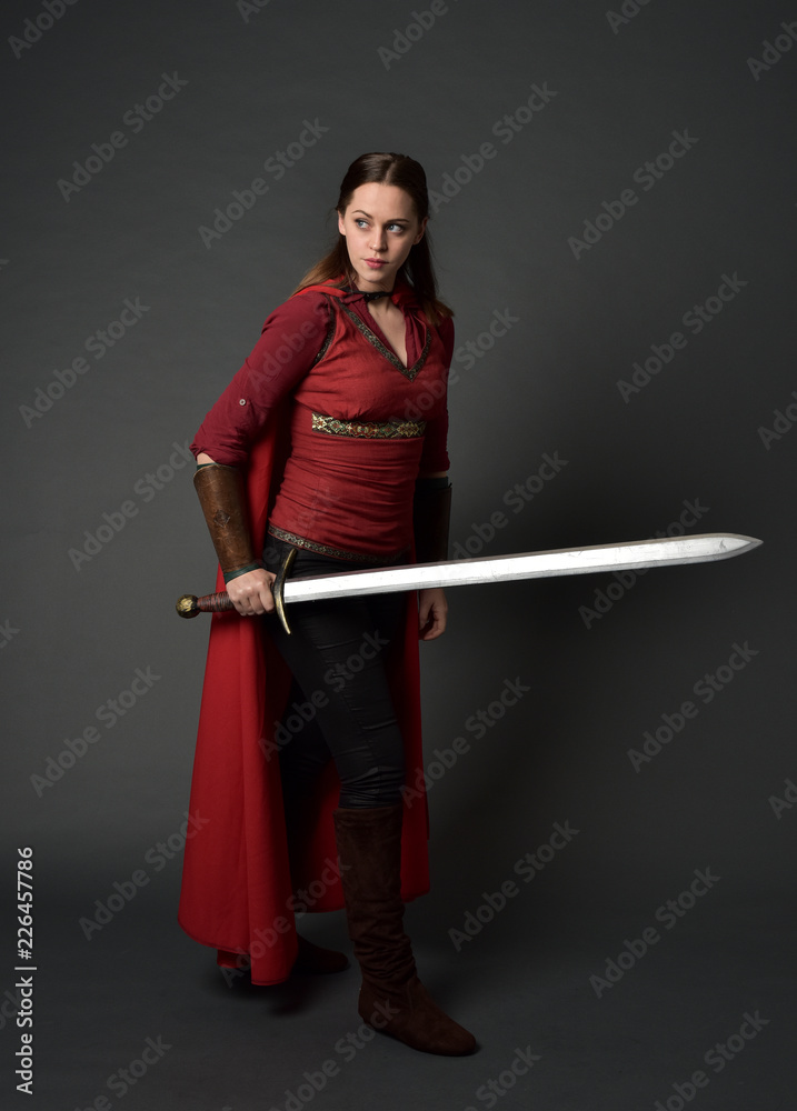 full length portrait of brunette girl wearing red medieval costume and cloak. standing pose holding a sword on grey studio background. full length portrait of brunette girl wearing red medieval costume and cloak. standing pose holding a sword on grey studio background.