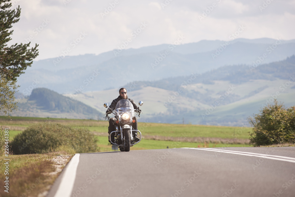 Motorcyclist in black leather outfit riding cruiser bike along road on bright sunny summer day on background foggy distant green woody hills under blue morning sky.