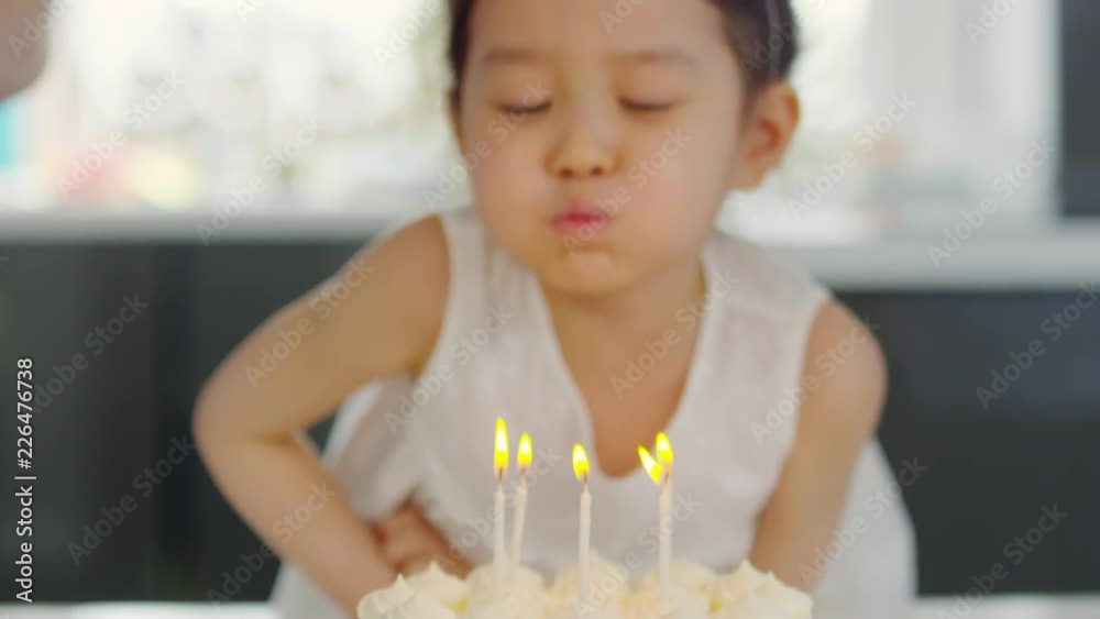Dolly shot of cute Asian girl in party hat blowing out candle on birthday cake as unrecognizable family clapping and celebrating