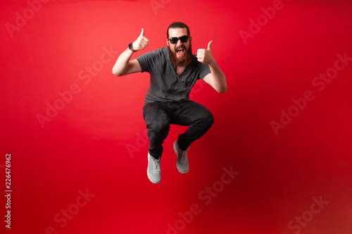 Obraz na płótnie Cheerful bearded hipster man with sunglasses jump over red background and showin
