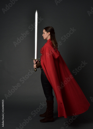  full length portrait of brunette girl wearing red medieval costume and cloak. standing pose  holding a sword on grey studio background. photo