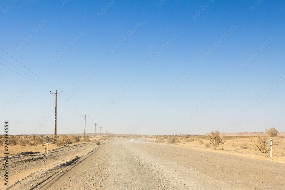 Dusty desert road in the Maranjab desert, in Iran, surrounded by sand and dry bushes, with the shape of a car visible in the background