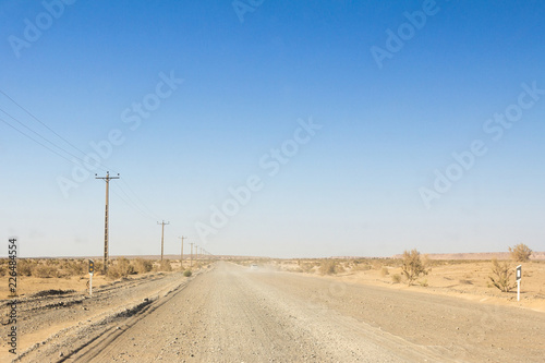 Dusty desert road in the Maranjab desert, in Iran, surrounded by sand and dry bushes, with the shape of a car visible in the background © Jerome