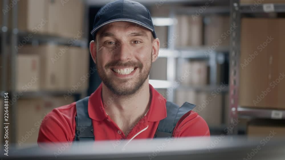 Portrait of Uniformed Worker Using Personal Computer while Sitting at His Desk in the Warehouse. In the Background, Room with Shelves Full of Cardboard Box Packages Ready For Shipping. Portrait of Uniformed Worker Using Personal Computer while Sitting at His Desk in the Warehouse. In the Background, Room with Shelves Full of Cardboard Box Packages Ready For Shipping.