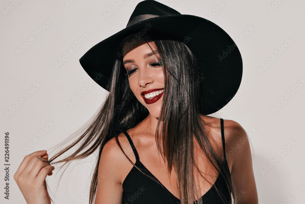 Studio shot of wonderful young woman with bright make up and dark hair wearing black hat and dress posing on isolated background with wonderful smile and closed eyes, holiday, Halloween Studio shot of wonderful young woman with bright make up and dark hair wearing black hat and dress posing on isolated background with wonderful smile and closed eyes, holiday, Halloween