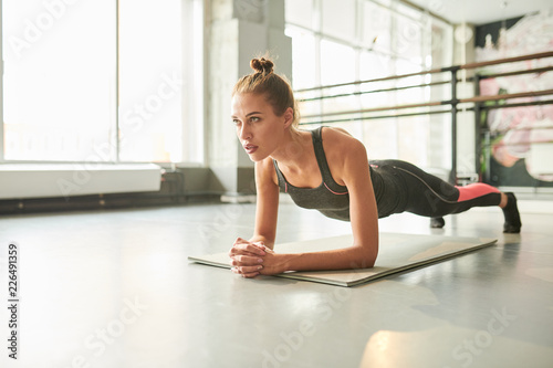 Full length portrait of contemporary young woman doing plank exercise during fitness workout in health club, copy space