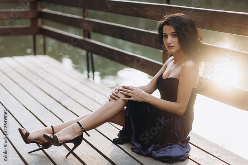 Stunning young woman in seductive long black dress with naked shoulders sits on the bridge over a lake in the rays of evening sun