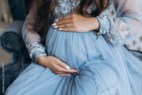 Sensetive portrait of pregnant woman. Beautiful expecting woman in blue dress poses in a studio with luxury design