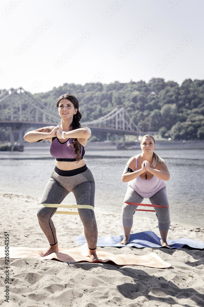 Productive workout. Happy positive women doing sit ups while using elastic band for training Productive workout. Happy positive women doing sit ups while using elastic band for training