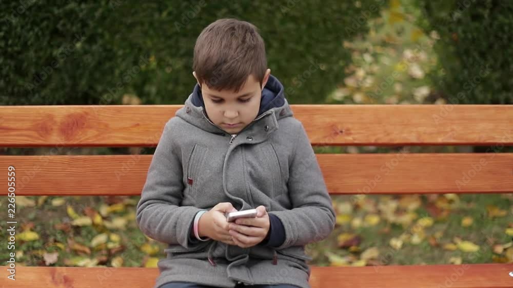 Little boy use a phone in park. He sits on the bench in autumn weather. Yellow trees on background Little boy use a phone in park. He sits on the bench in autumn weather. Yellow trees on background