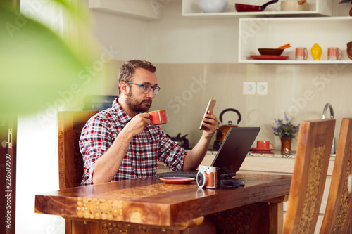 Modern bearded man using cellphone and drinking coffee with laptop.