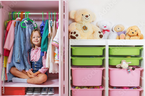 A little child girl is sitting in a wardrobe in a children's room. Saving and storage system for children's things and toys concept.