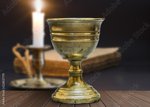 Old chalice on wooden table next to a candle and an closed book