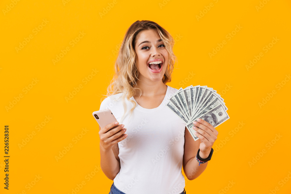 Photo of pretty woman in basic clothing holding fan of dollar money, isolated over yellow background Photo of pretty woman in basic clothing holding fan of dollar money, isolated over yellow background