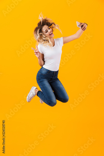 Full length image of young woman jumping and taking selfie photo on mobile phone, isolated over yellow background