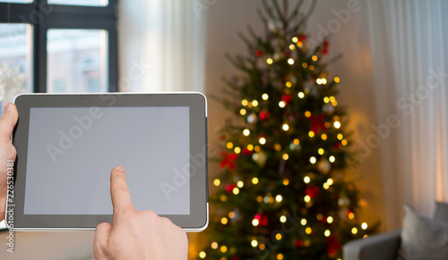 holidays and technology concept - close up of male hands with tablet pc over christmas tree lights background