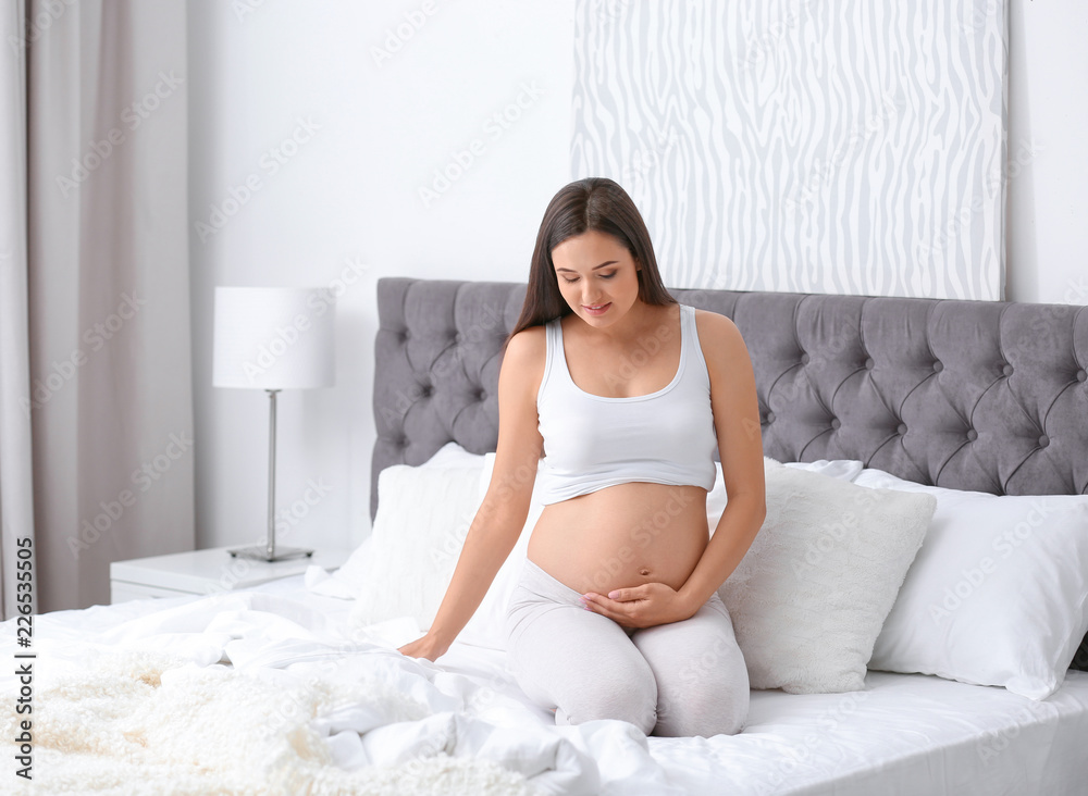 Young beautiful pregnant woman sitting on bed and touching her belly at home Young beautiful pregnant woman sitting on bed and touching her belly at home