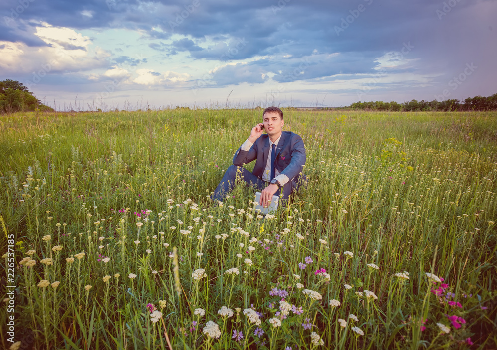 Businessman sitting in his suit on a green sunny field phoning from his mobile phone