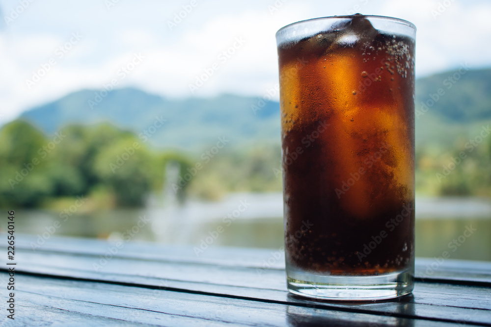 Cola in glass on table with mountain and river background Cola in glass on table with mountain and river background