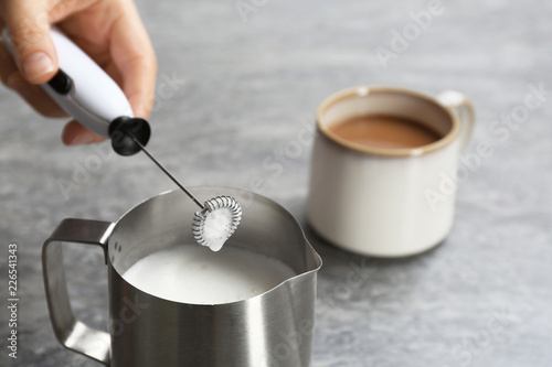 Woman using milk frother in pitcher near cup of coffee on table photo