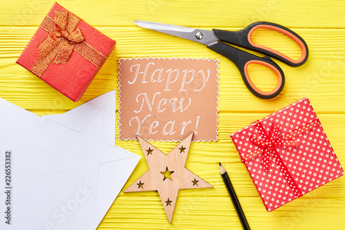 Happy New Year wooden background. Paper note with text, red gift boxes, cut out wooden Christmas star, scissors on yellow wooden background.